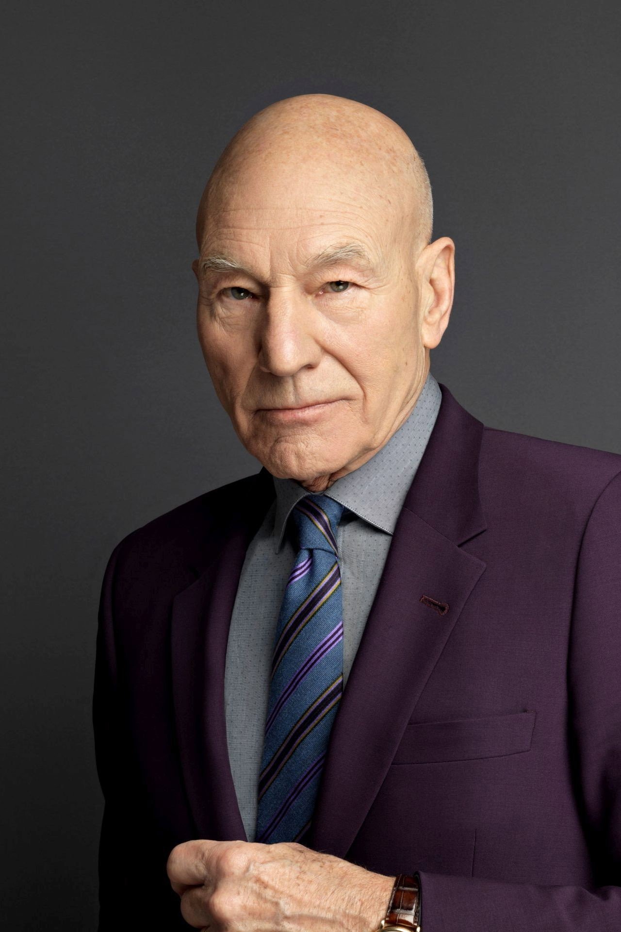 Patrick Stewart con traje morado y corbata de rayas azules frente a un fondo gris, mirando a cámara