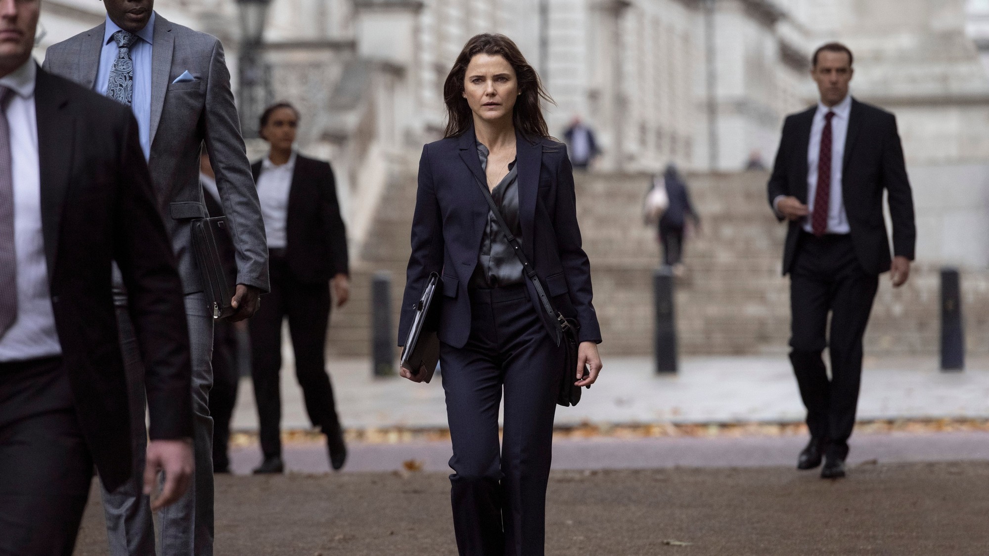 Keri Russell como Kate Wyler camina con traje oscuro y un portafolios por una calle de Londres, rodeada de escoltas y asesores