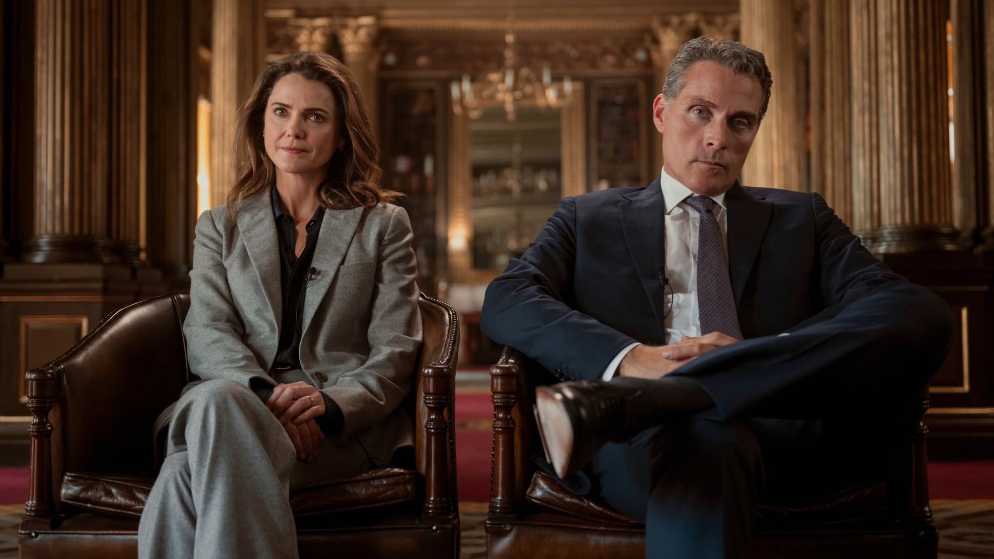 Kate y Hal Wyler sentados en sillones de cuero en un salón con columnas doradas, mirando de frente con gesto serio