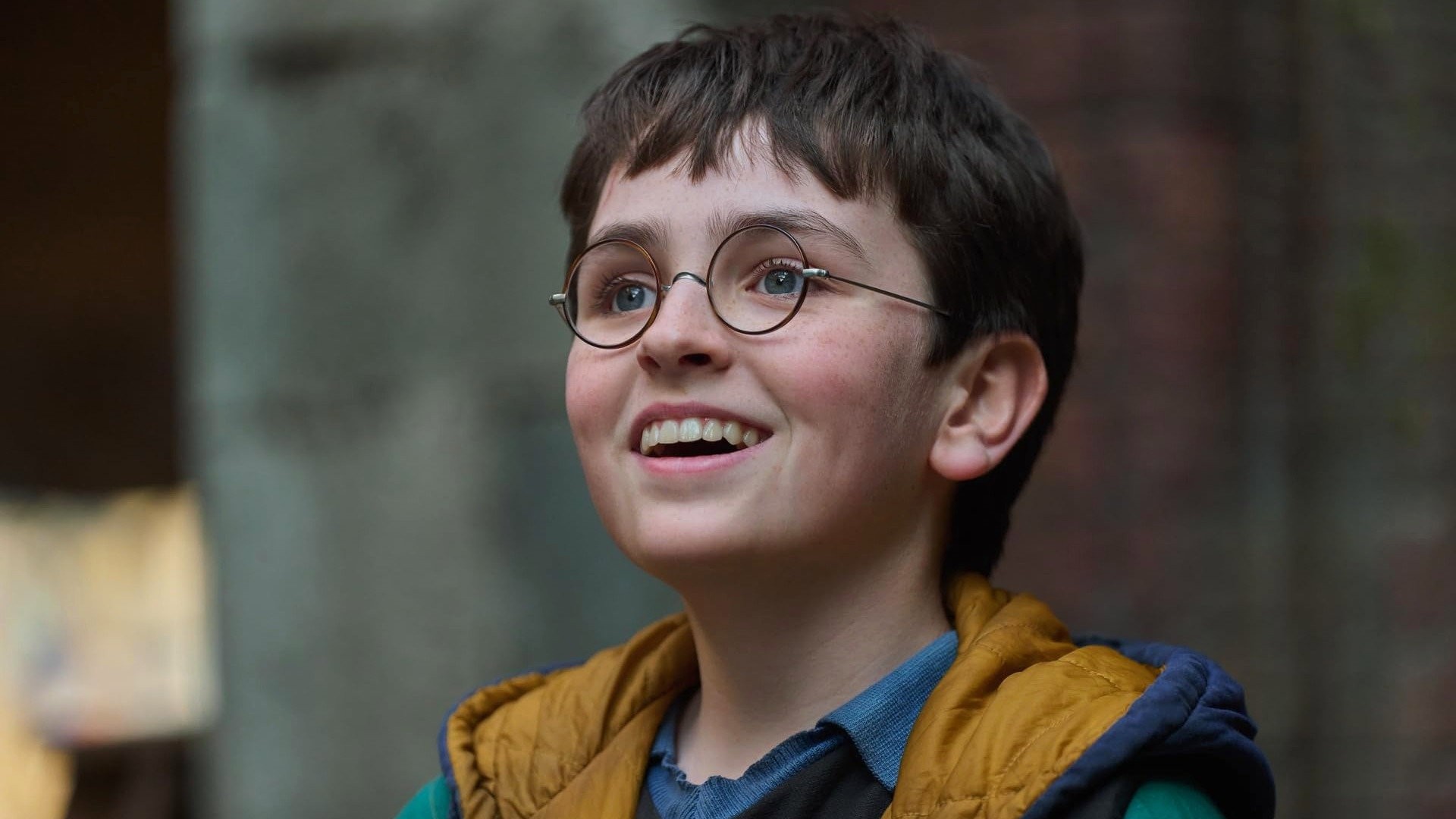 Harry Potter, con sus gafas redondas y un chaleco acolchado color mostaza, sonríe mirando hacia arriba
