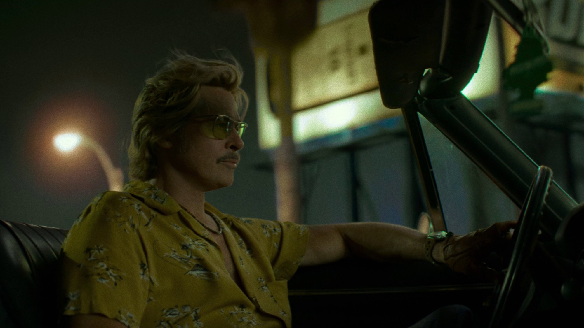 Cliff Booth, con camisa amarilla estampada y gafas de sol, al volante de un auto descapotable bajo las luces de la calle