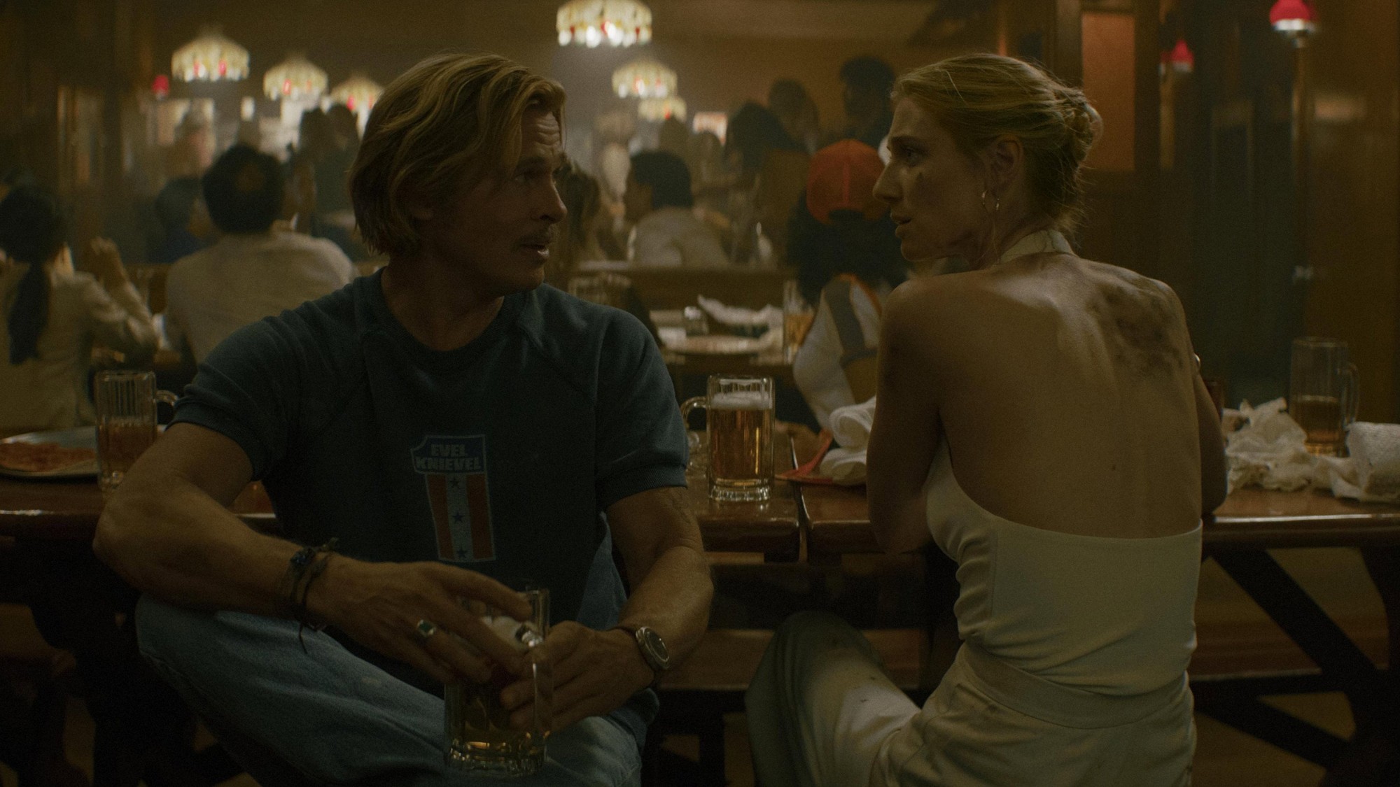 Cliff Booth, con camiseta azul y un tarro de cerveza, conversa con una mujer de vestido blanco en un bar concurrido